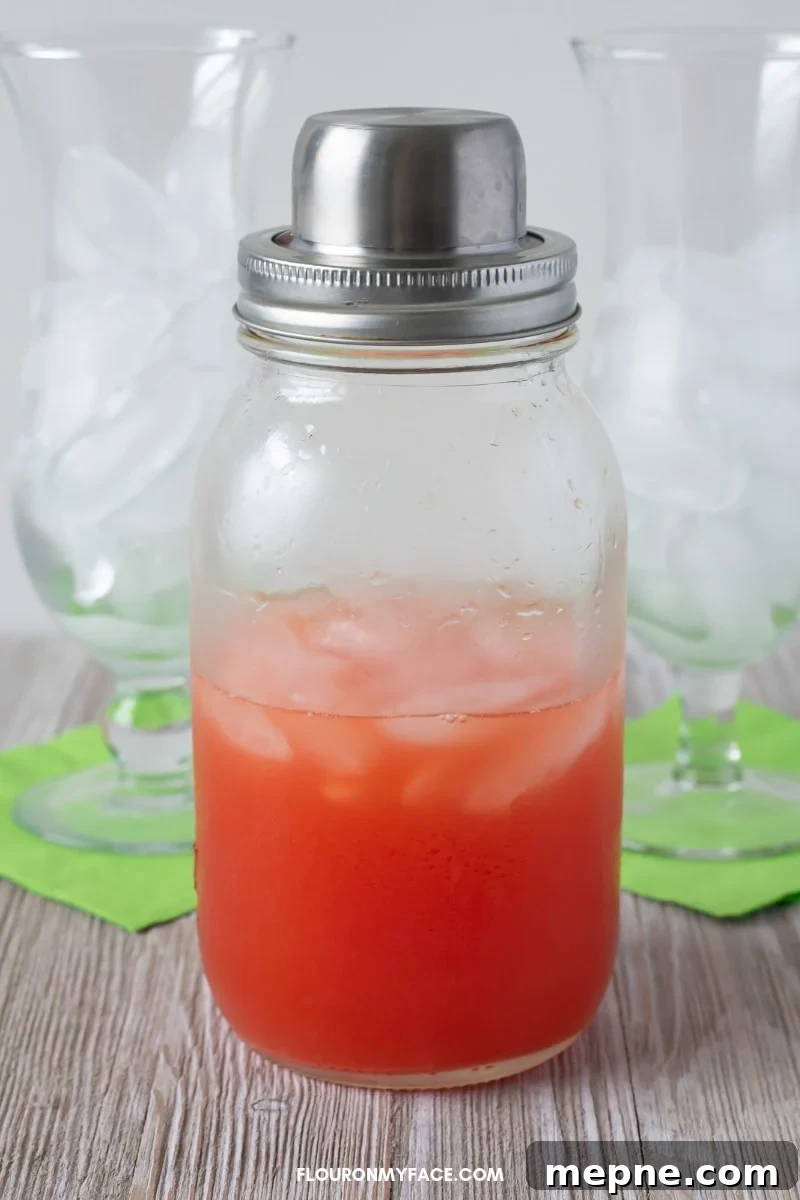 Classic Fruity Hurricane Cocktail Recipe 5 Close up photo of a clear Mason jar cocktail shaker filled with a classic Hurricane with two hurricane cocktail glasses in the background.