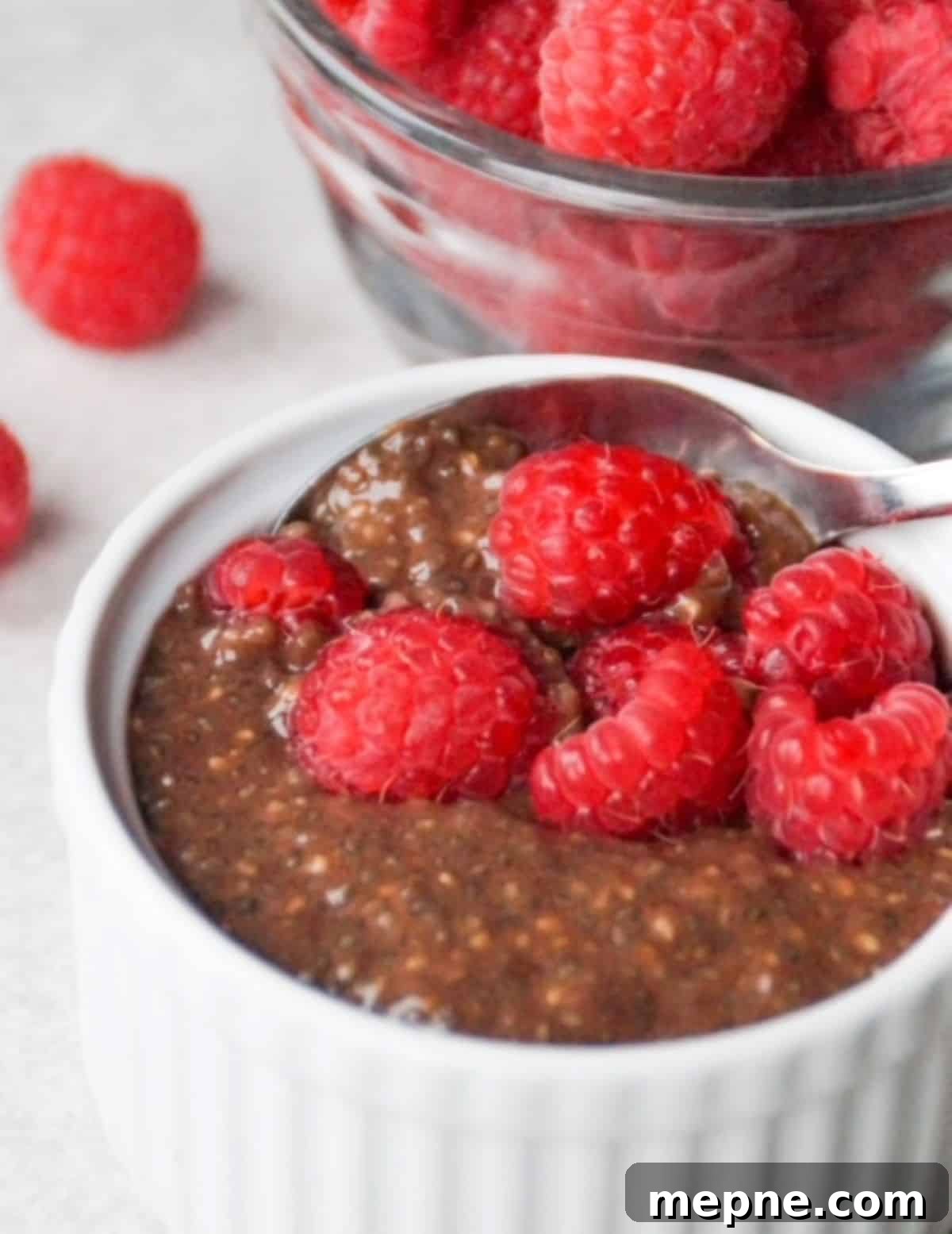 Chocolate chia pudding in a small white ramekin with a spoon and topped with fresh raspberries. One small glass bowl with fresh raspberries is behind the white ramekin.