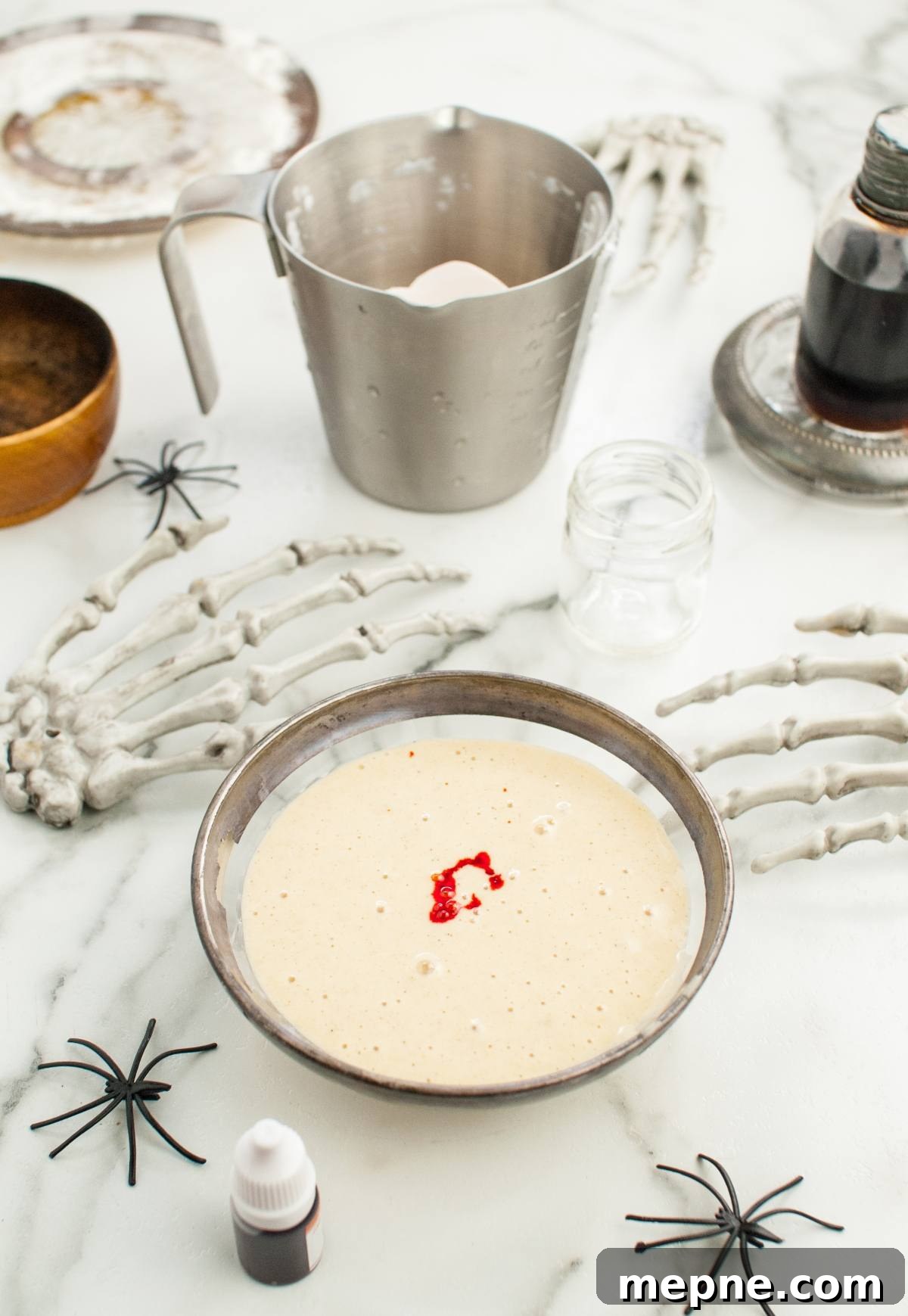 Bowl of batter with red food coloring, surrounded by skeleton hands and Halloween decorations on a marble surface—perfect for making Colorful Halloween Pancakes or trying fun Halloween recipes for kids.