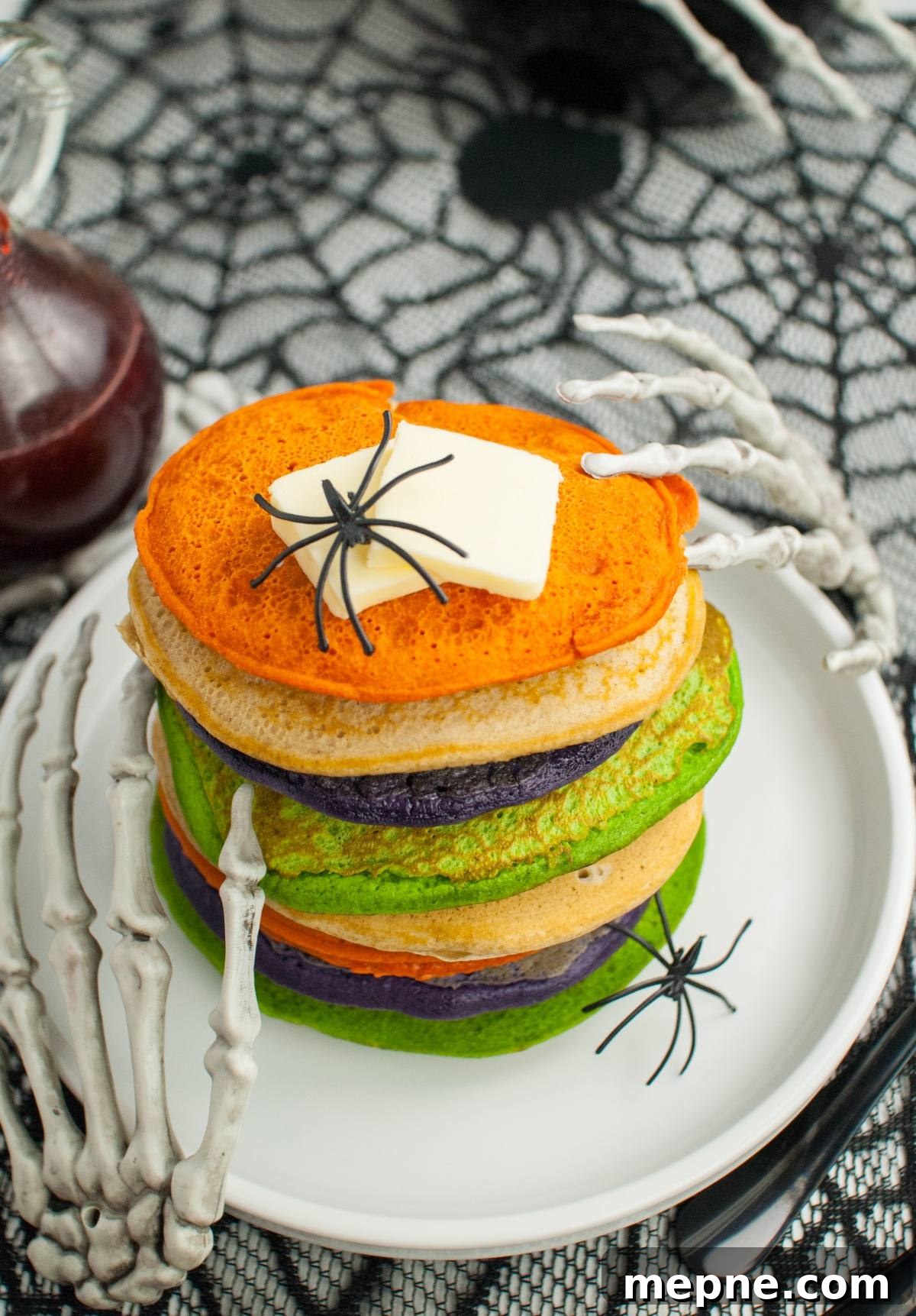Stack of colorful Halloween pancakes topped with butter, fake spiders, and skeleton hands on a spiderweb tablecloth—perfect for spooky breakfast ideas or Halloween party food!.