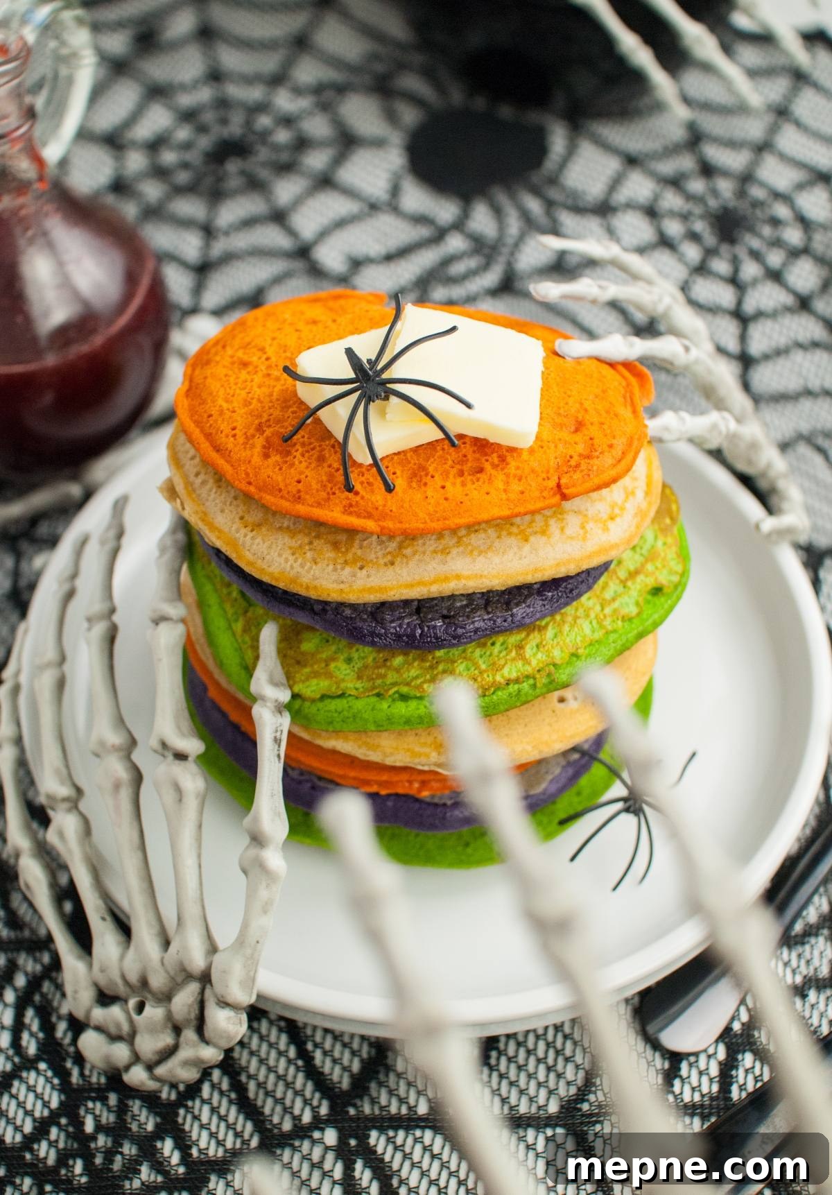 Stack of colorful pancakes, topped with butter and a fake spider, held by skeleton hands—perfect for spooky breakfast ideas or as fun Halloween party food.