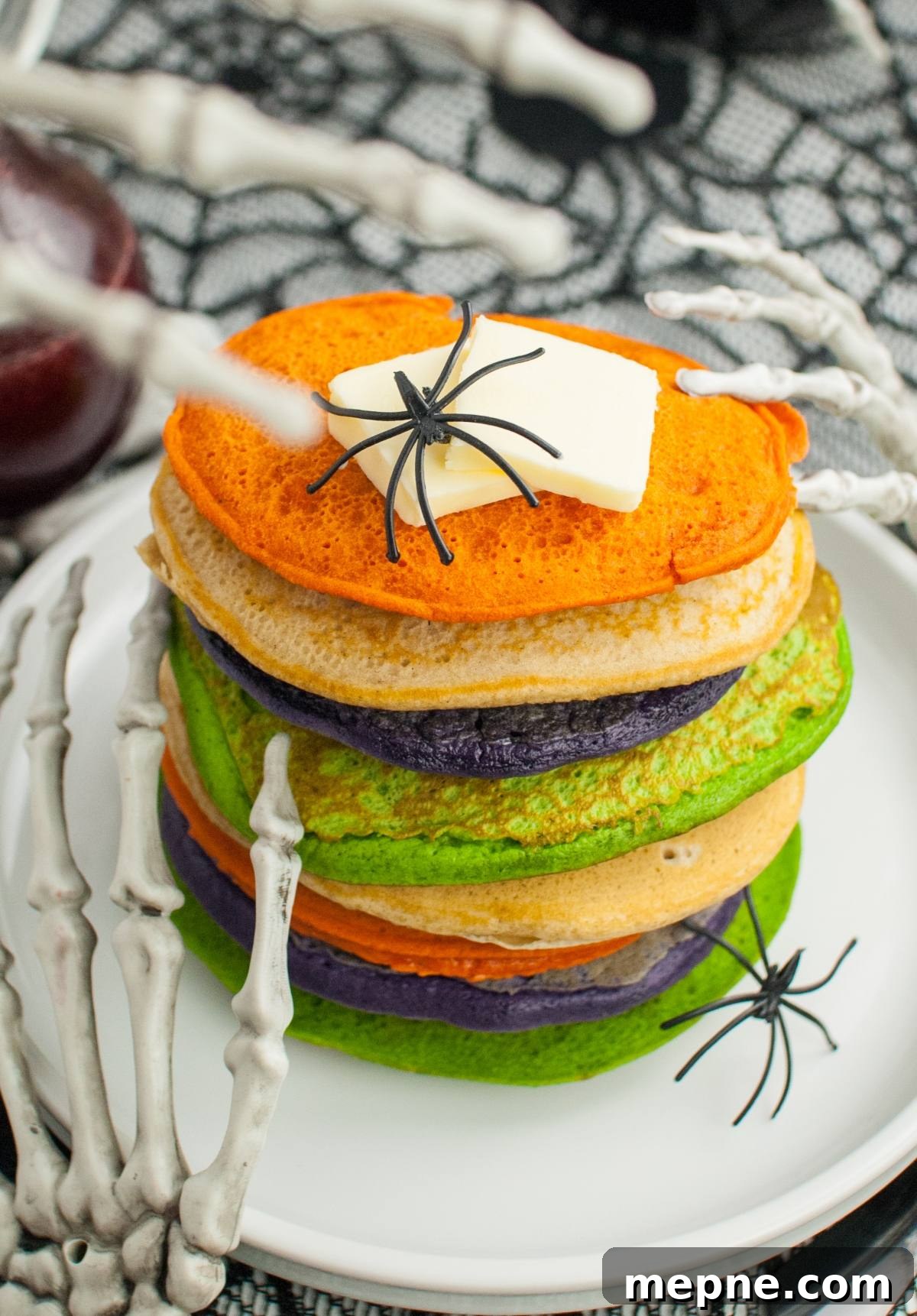 Stack of colorful pancakes with fake spiders, butter on top, and skeleton hands holding the plate—perfect Halloween party food for kids who love spooky treats!.