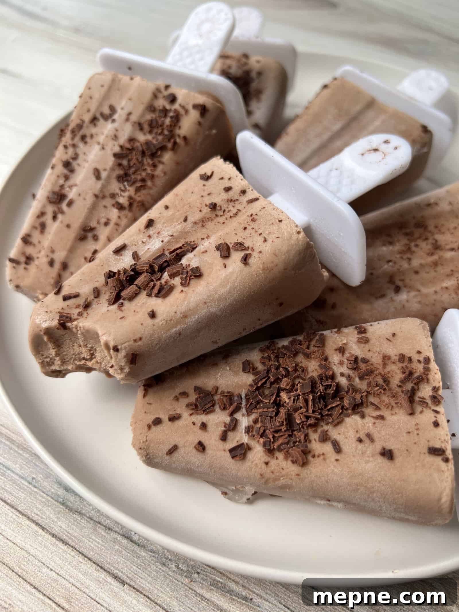 Homemade Fudgesicle Recipe: Creamy Chocolate Pops 2 Several creamy chocolate popsicles with plastic handles are arranged on a plate. Some have chocolate shavings sprinkled on top, and one popsicle has a bite taken out of it. The background is a light wooden surface.