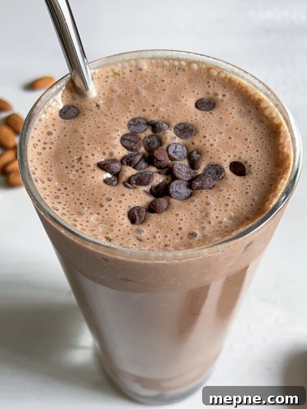 A glass of chocolate smoothie topped with chocolate chips, served with a metal straw. Some whole almonds are scattered in the background on a light surface.