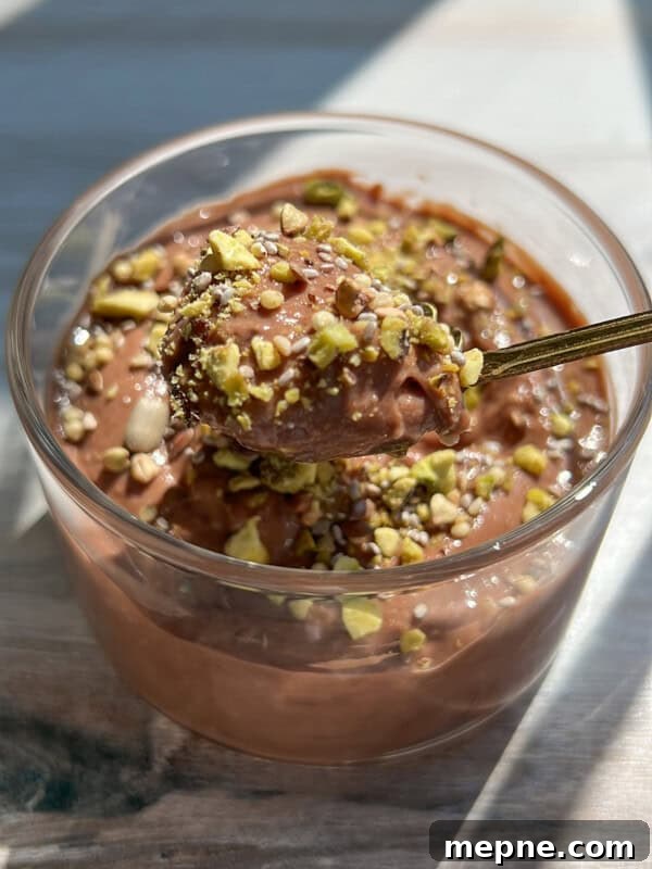 A glass cup filled with creamy chocolate mousse, topped with chopped pistachios. A spoon is lifting a portion of the mousse, showing its smooth texture. Sunlight illuminates the dessert on a wooden surface.