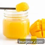 A spoonful of mango jelly set across the top of a jar of mango jelly next to some fresh cut mango.