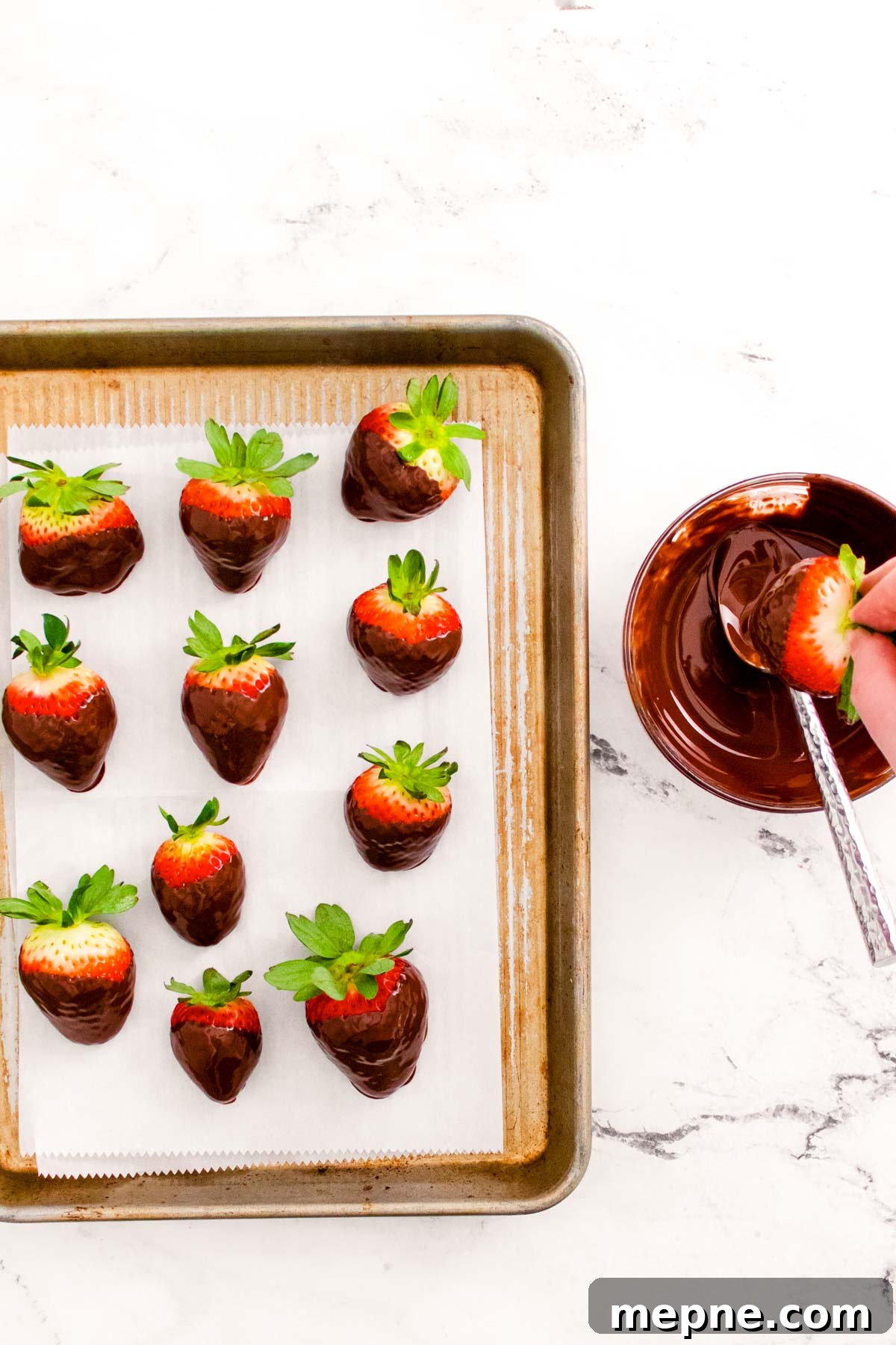 Chocolate covered strawberries on a baking sheet and a woman's hand dipping more in chocolate.