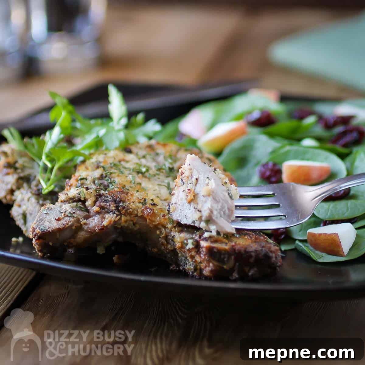 Affordable Meats to Buy and Cuts You Should Avoid 6 Side view of blue cheese encrusted pork chops garnished with herbs with a side of spinach sprinkled with apples and cranberries on a black plate.