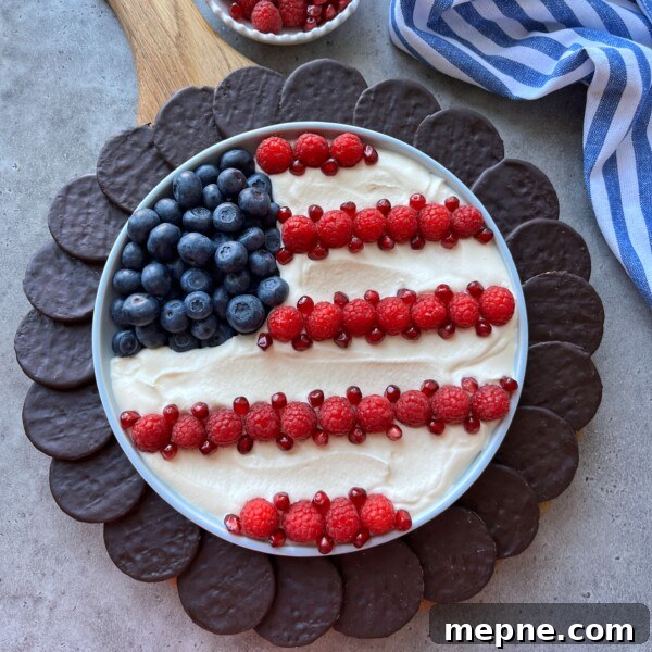 Red White and Blue Caprese Bites for Fourth of July 6 A round 4th of July dessert dip platter decorated like the American flag with blueberries, raspberries, and pomegranate seeds on a creamy base, surrounded by chocolate cookies. A striped blue and white cloth is nearby.