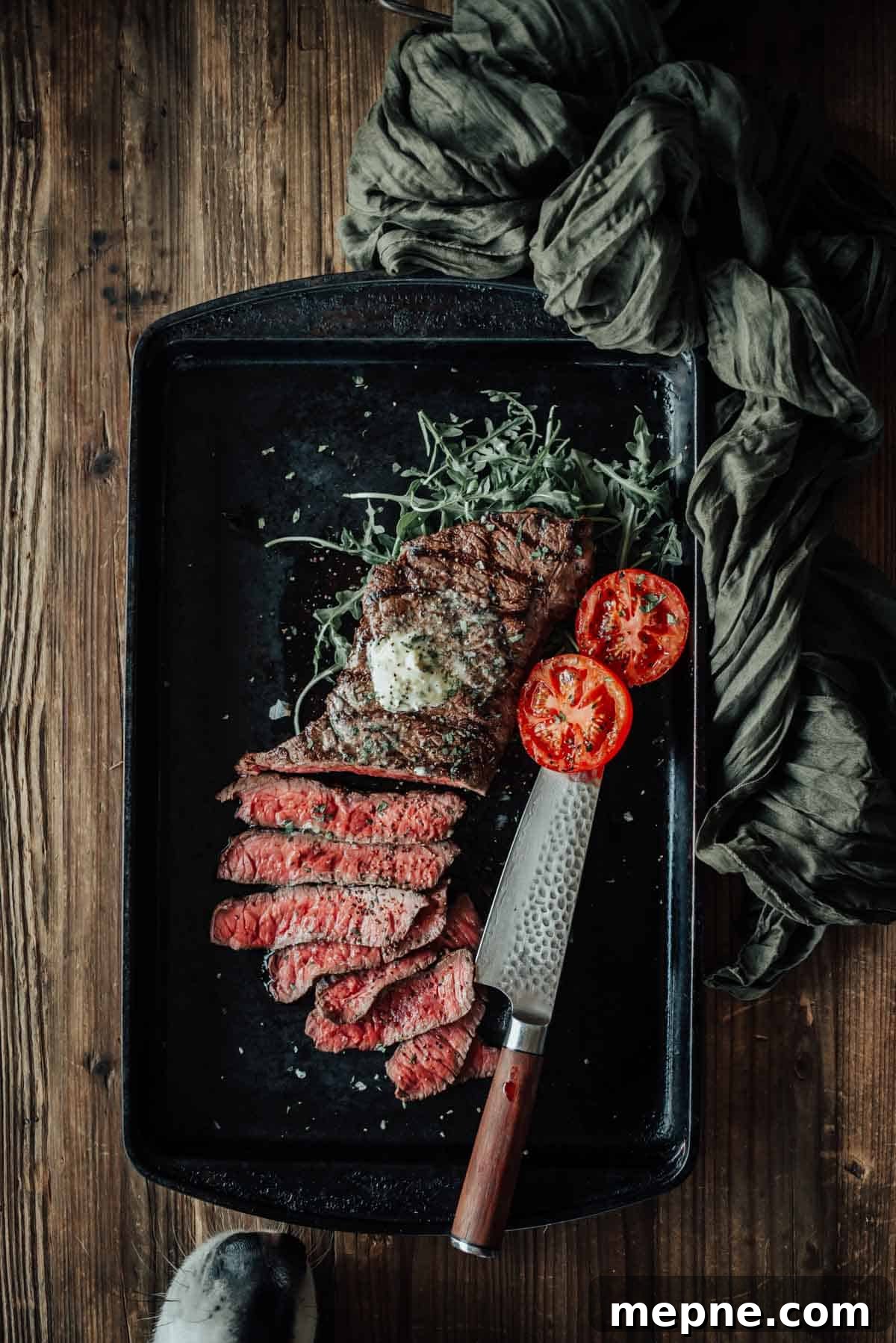 How to Grill London Broil for Tender Juicy Results 2 A grilled london broil with half sliced into pieces, garnished with a pat of butter, vegetables, and two grilled tomato halves, served on a black tray with a knife and a dogs nose sneaking in near the edge for a sniff.