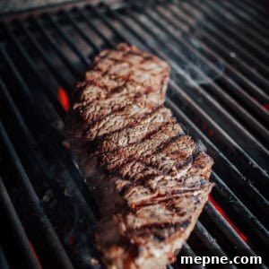How to Grill London Broil for Tender Juicy Results 13 A london broil being grilled on an open flame grill, with visible char marks and red-hot coals underneath.