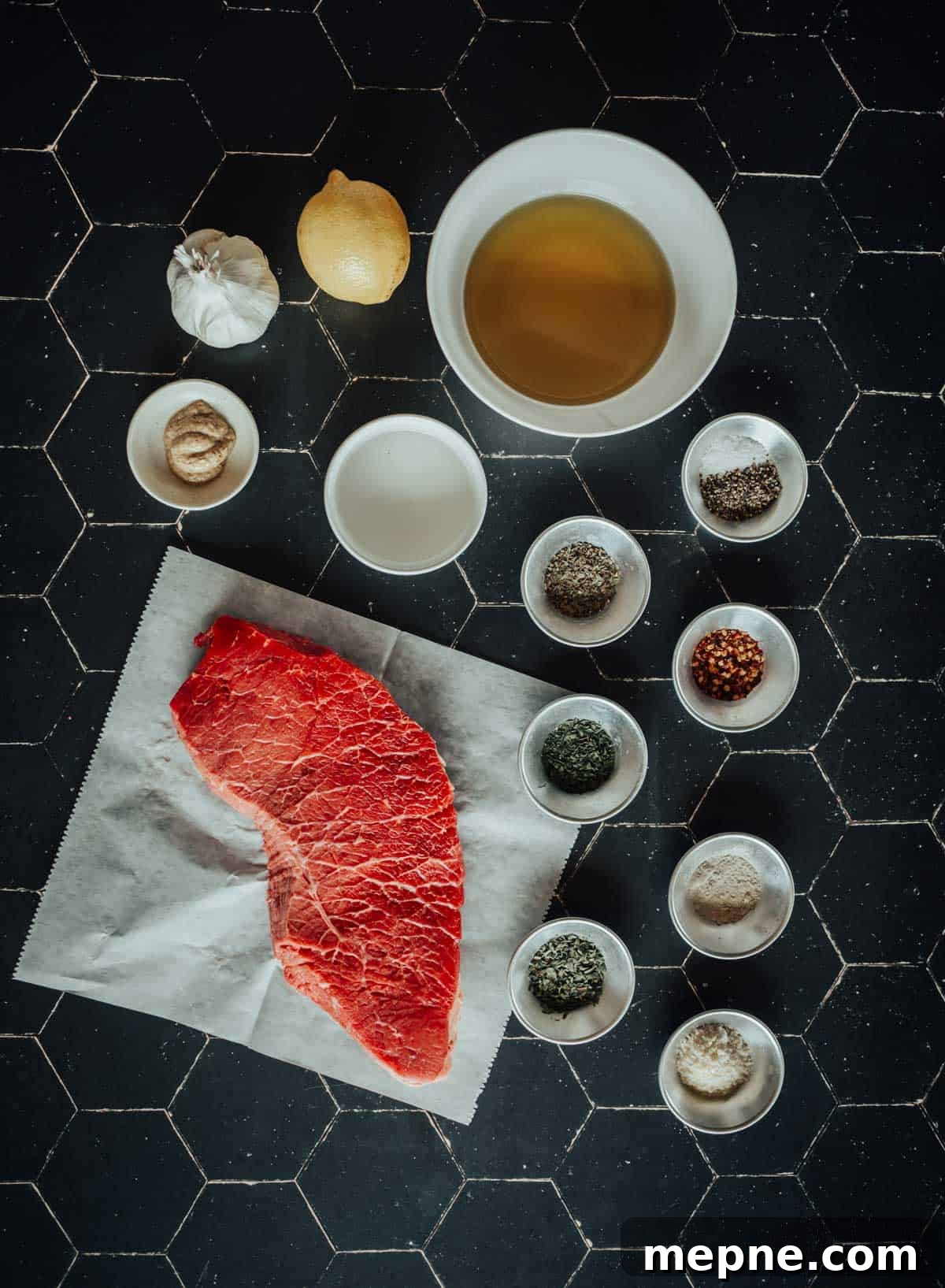 How to Grill London Broil for Tender Juicy Results 4 Raw london broil on parchment paper surrounded by bowls of spices, a garlic bulb, a lemon, and a bowl of oil, all placed on a black surface.