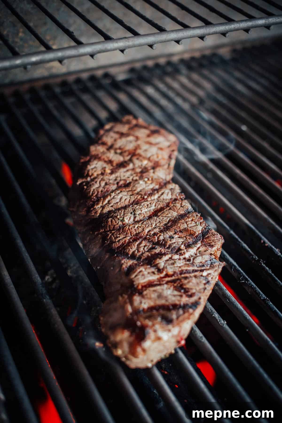 How to Grill London Broil for Tender Juicy Results 5 A london broil being grilled on an open flame grill, with visible char marks and red-hot coals underneath.