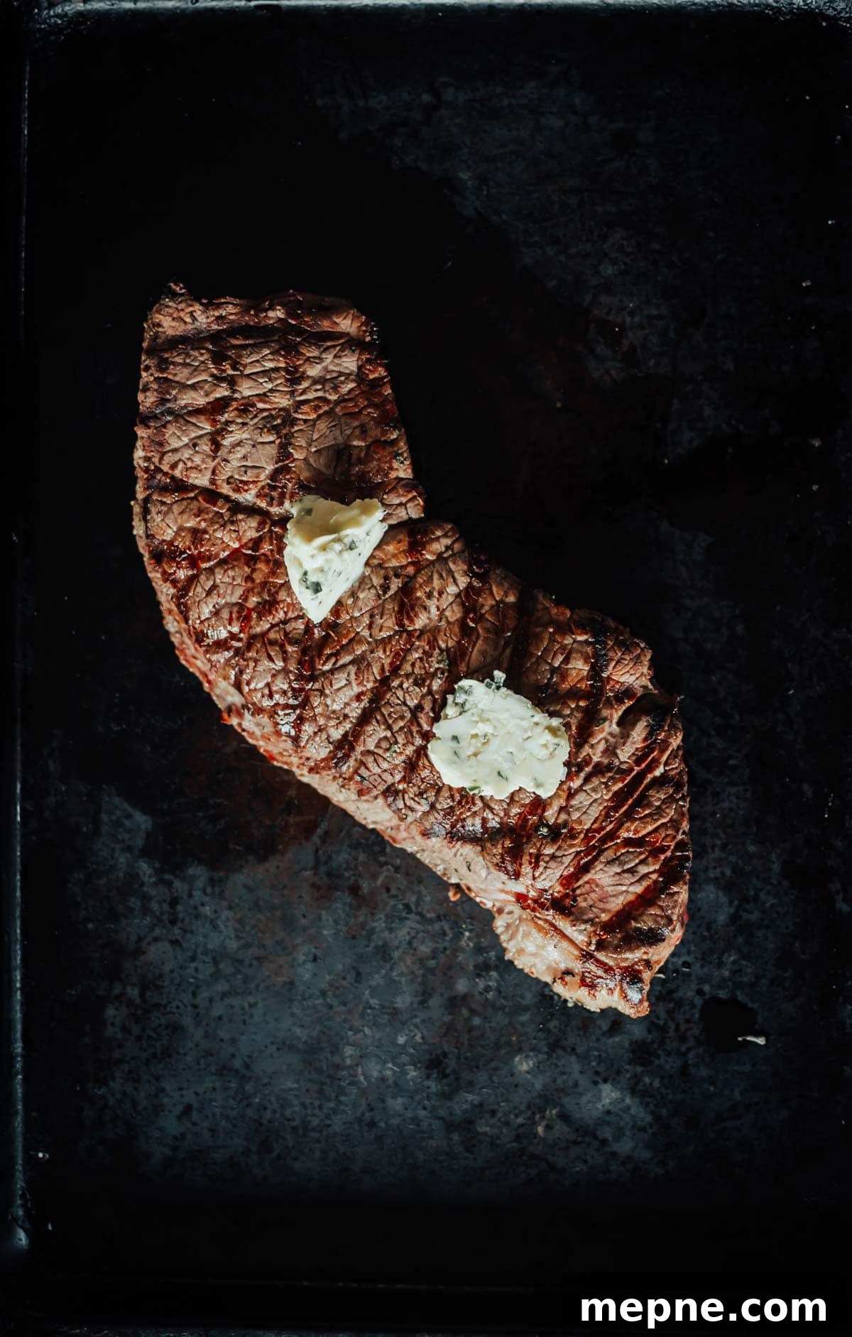 How to Grill London Broil for Tender Juicy Results 6 A grilled steak resting with two small pats of melting butter on top, placed on a dark surface.