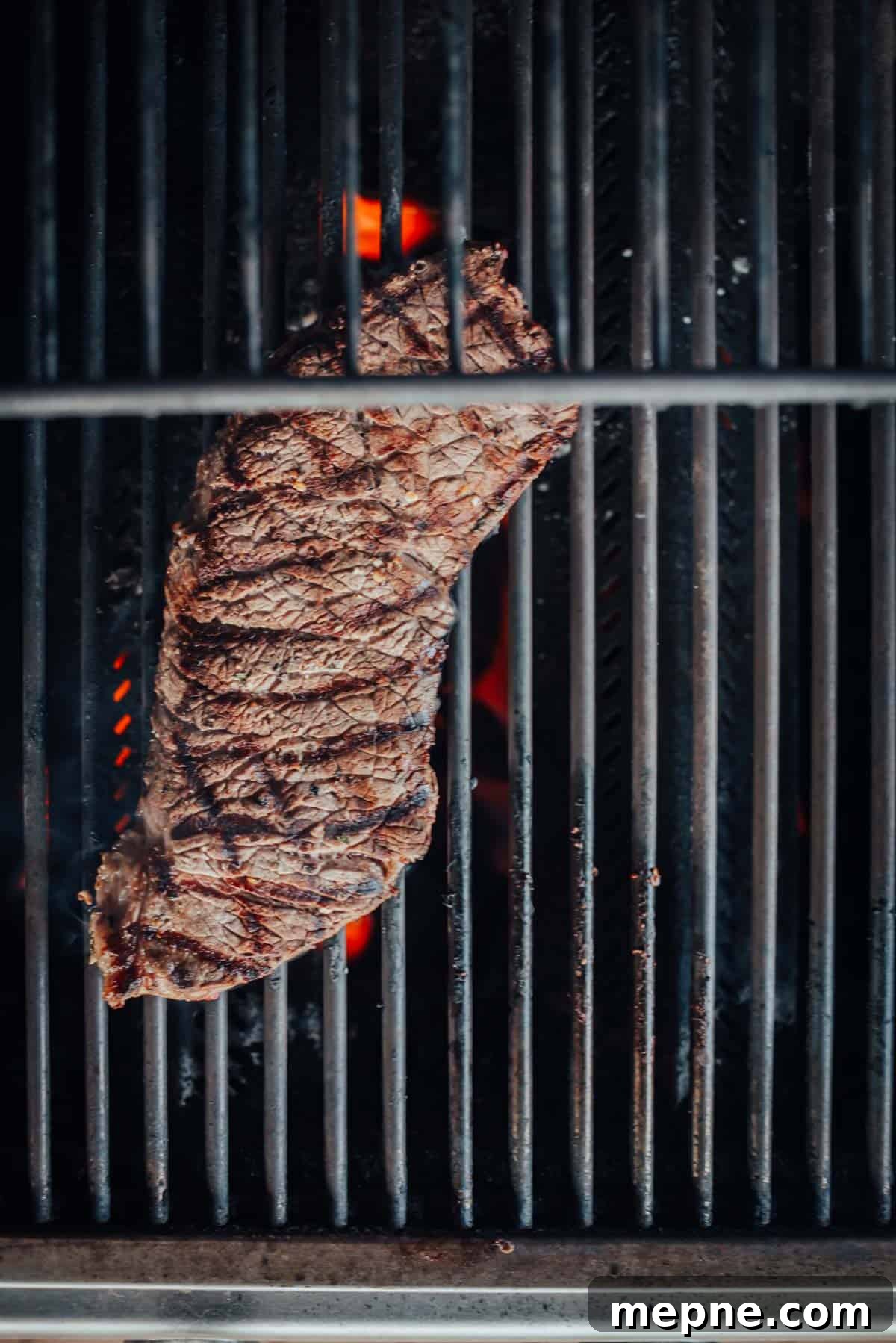 How to Grill London Broil for Tender Juicy Results 7 A london broil is being grilled on a barbecue, showing grill marks and flames beneath the grates.