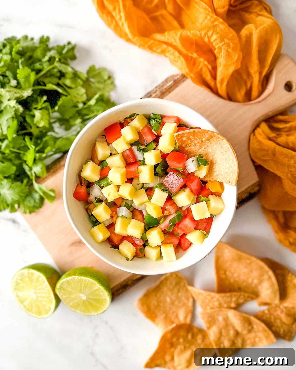 Pineapple Pico de Gallo Recipe: Tropical Salsa 2 Overhead shot of pineapple pico de gallo in a white bowl surrounded by tortilla chips, a halved lime, and fresh cilantro.