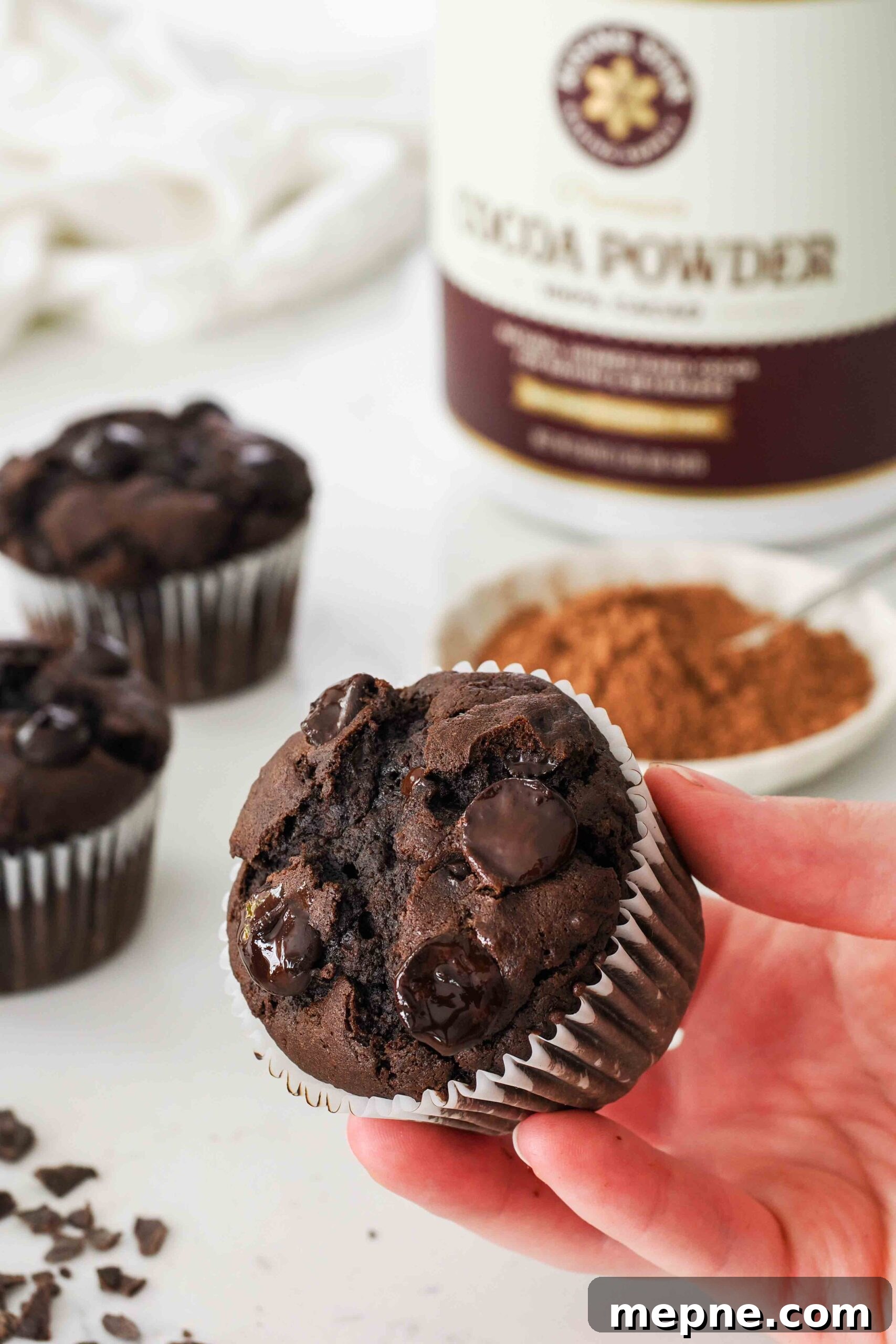 Moist and Fudgy Double Chocolate Muffins Recipe 2 A hand holds a double chocolate muffin in front of a container of cocoa powder.