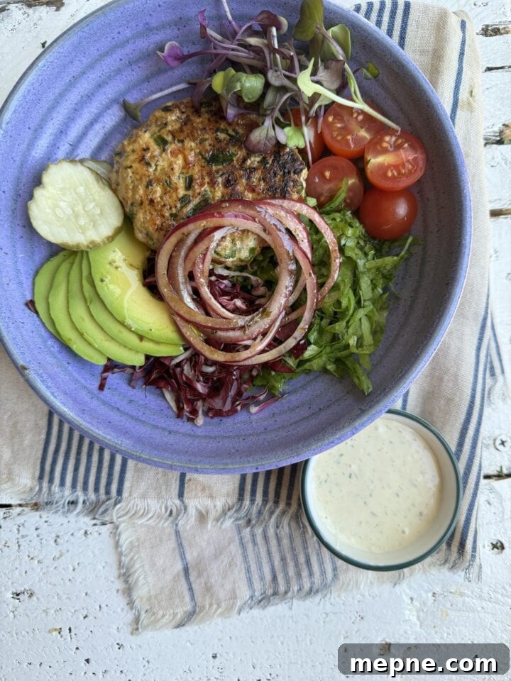 Smoky Chicken Patty Bowl: Weeknight Chicken Burger Recipe 13 blue bowl with chicken patty avocado cucumbers red onions white dressing and greens on a blue striped napkin with a small bowl with white sauce