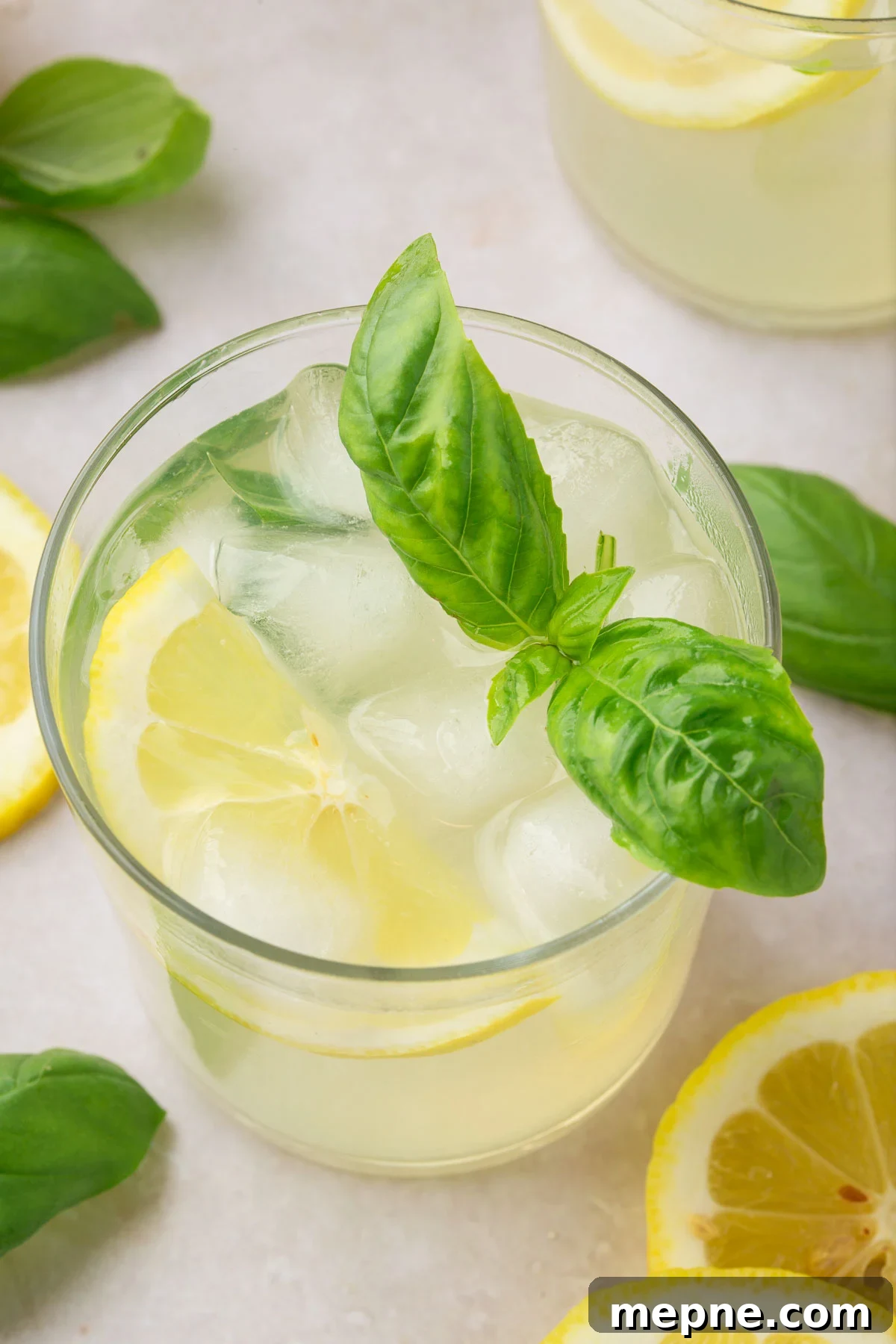 Fresh Basil Lemonade Recipe 10 A glass of basil lemonade filled with ice and topped with a slice of lemon and a few basil leaves with additional lemon slices and fresh basil surrounding the glass.